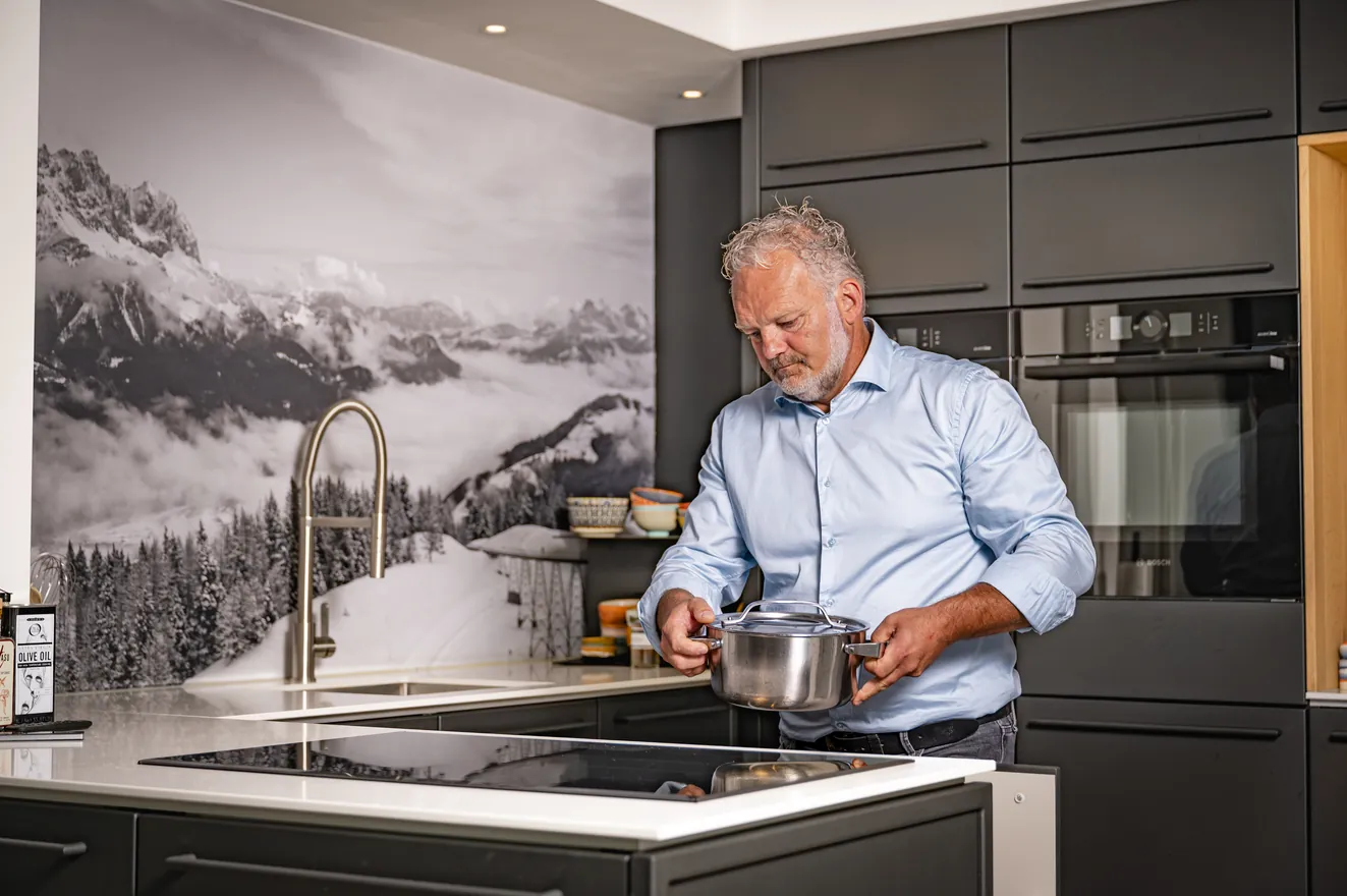 Henk van Veen kookt in een zwarte stalen keuken met een bergfoto als achterwand