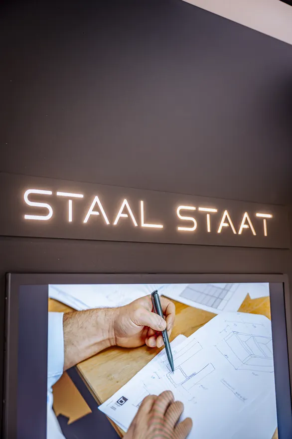 Het verlichte logo van Staal Staat boven een scherm met een tekening